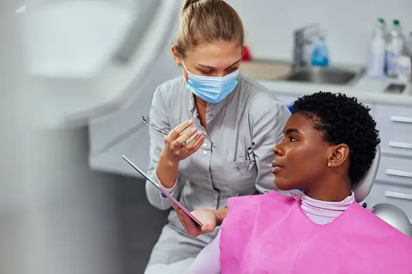 A dentist wearing a mask consulting a female patient, using a tablet to discuss her dental health during an examination.