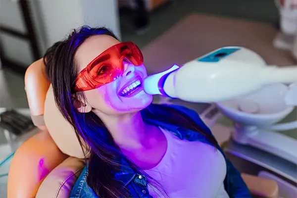 A woman in dental chair smiling brightly after a teeth whitening treatment, holding a mirror.